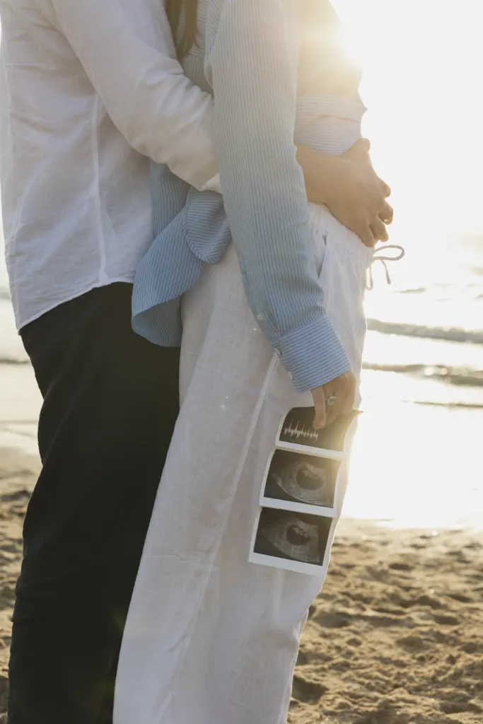 Aanstaande ouders aan zee met de echofoto's in de hand bij zonsondergang